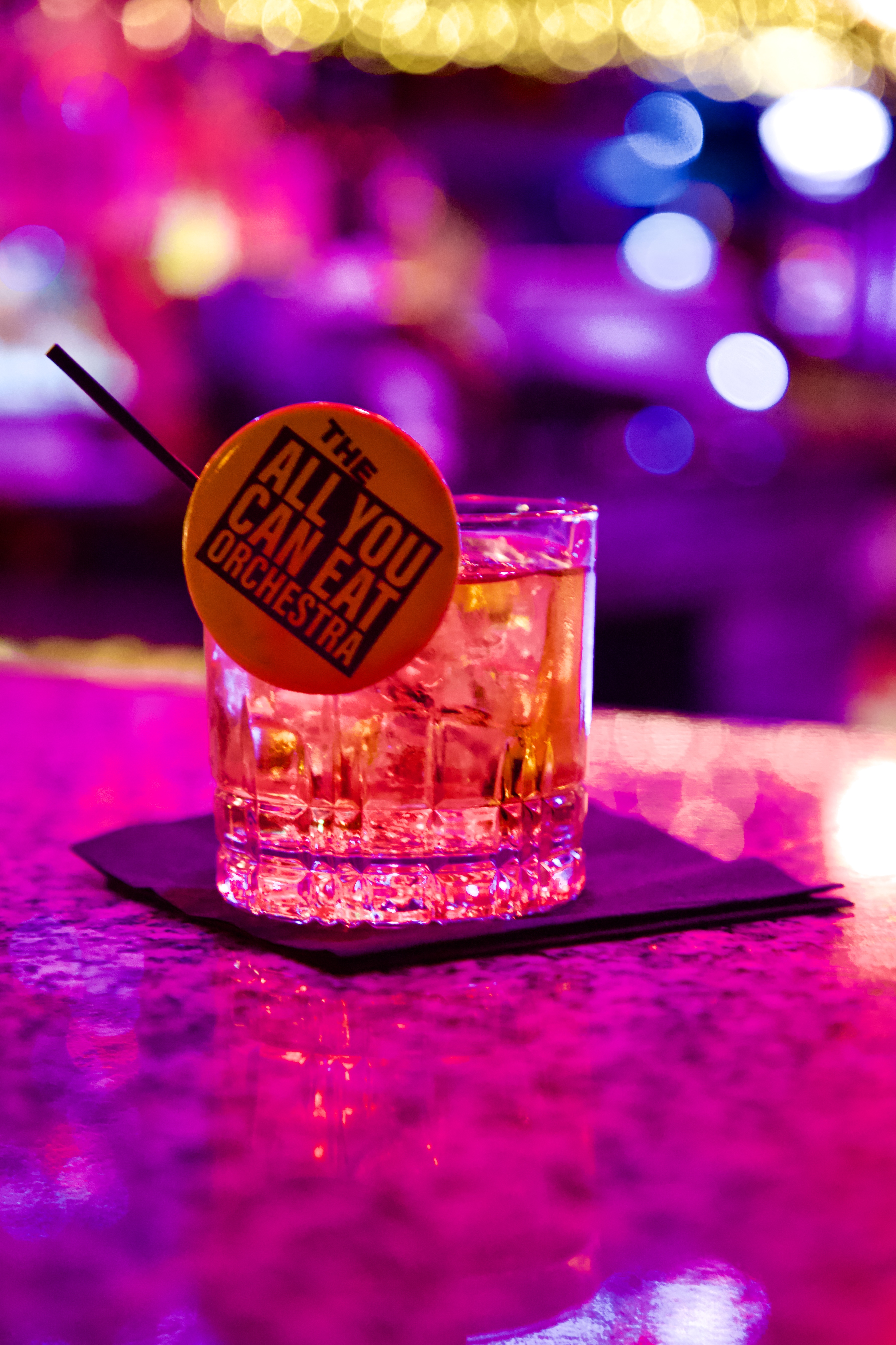 Branded cocktail garnished with a "The All You Can Eat Orchestra" badge on a neon-lit bar