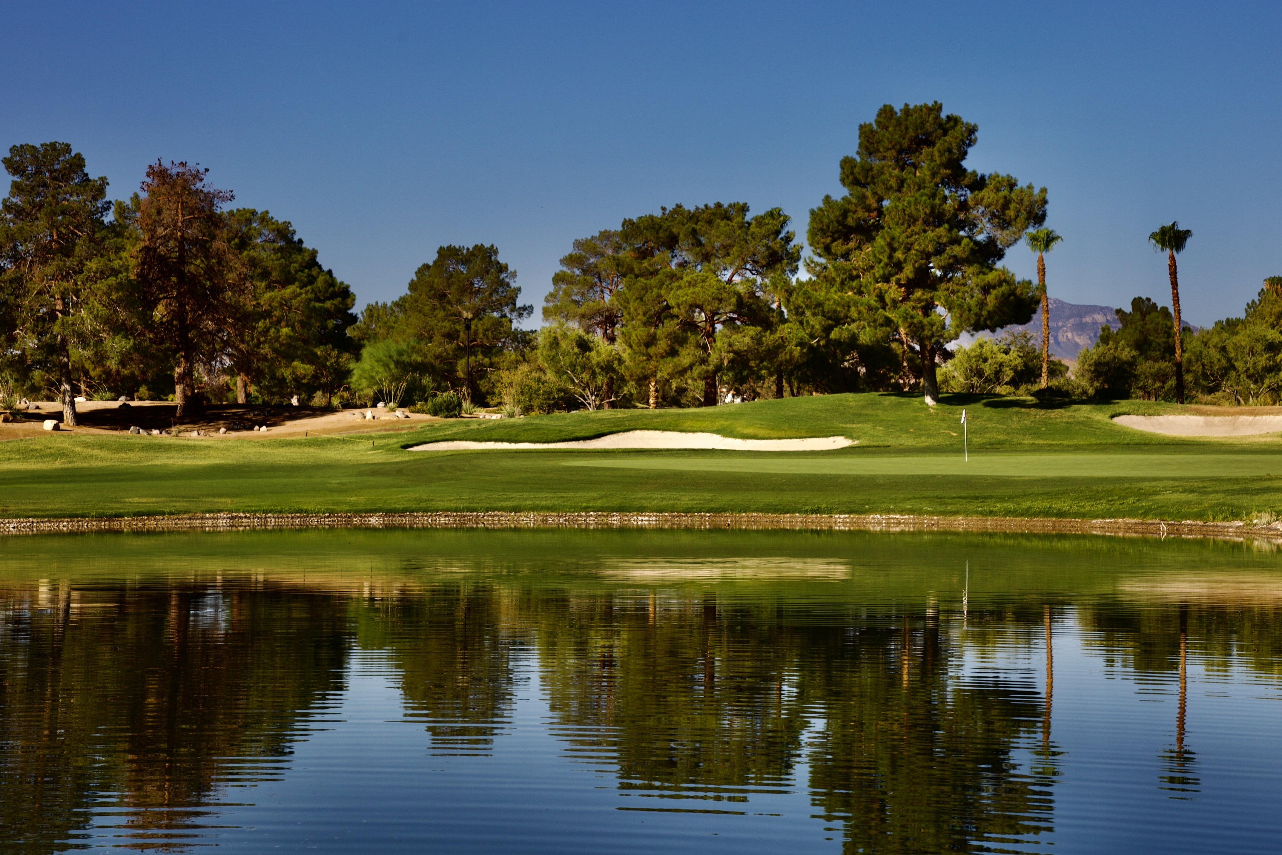 Golf green and flag mirrored across a glassy pond framed by pines and mountains