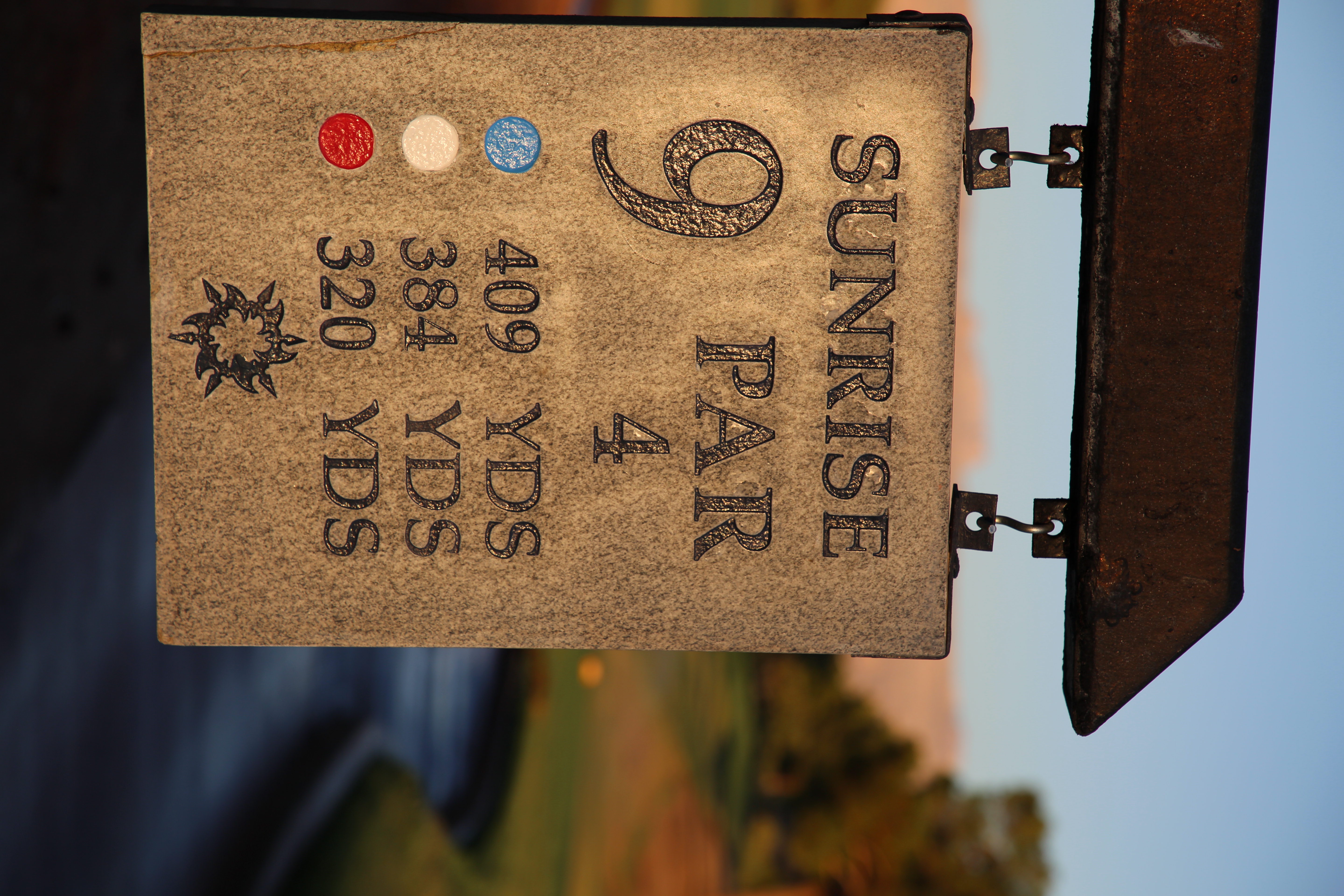 Engraved stone tee marker reading Sunrise Hole 9 Par 4 with yardages, hanging from an iron bracket