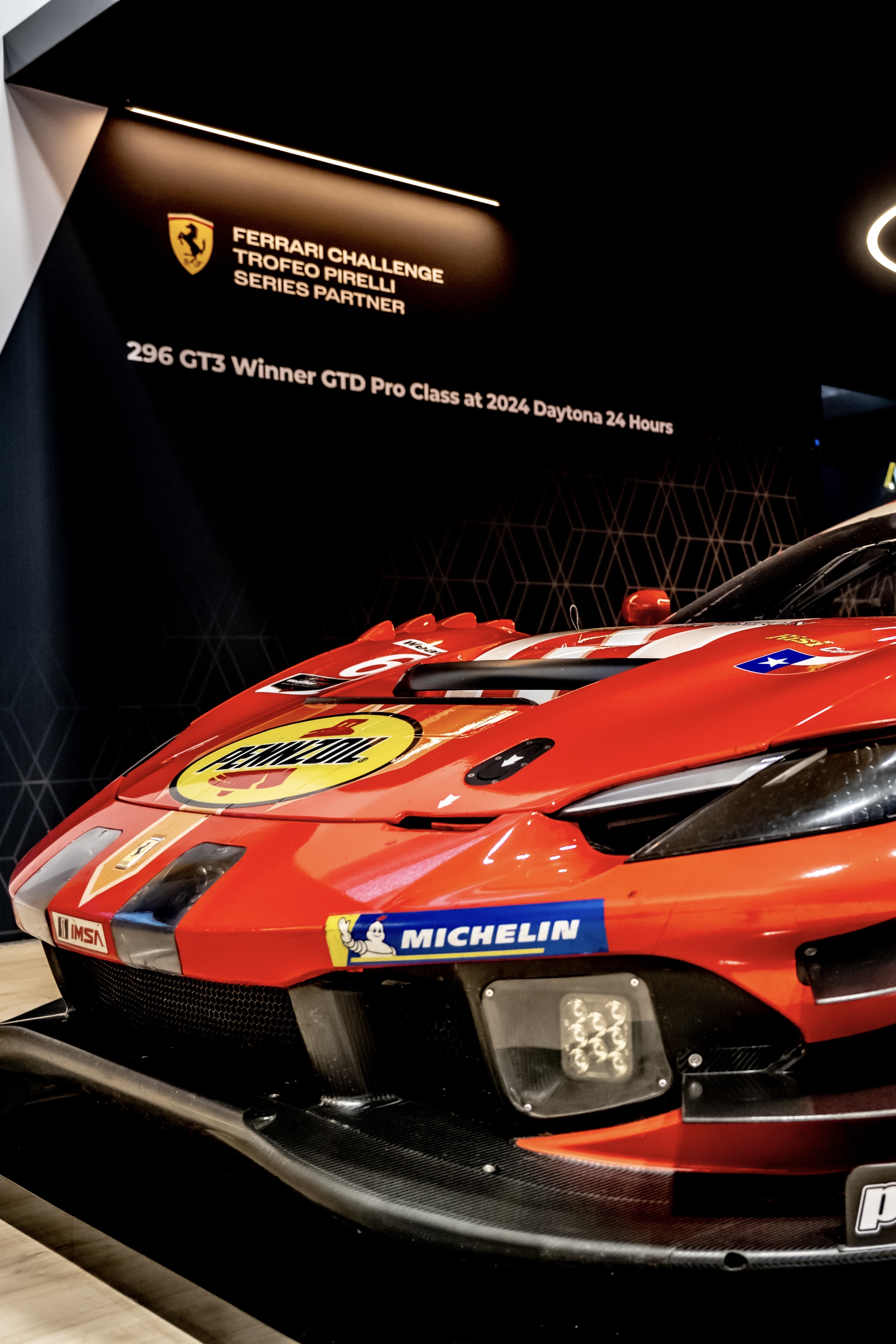 Front-quarter view of a red Ferrari 296 GT3 race car with Pennzoil and Michelin livery under event signage