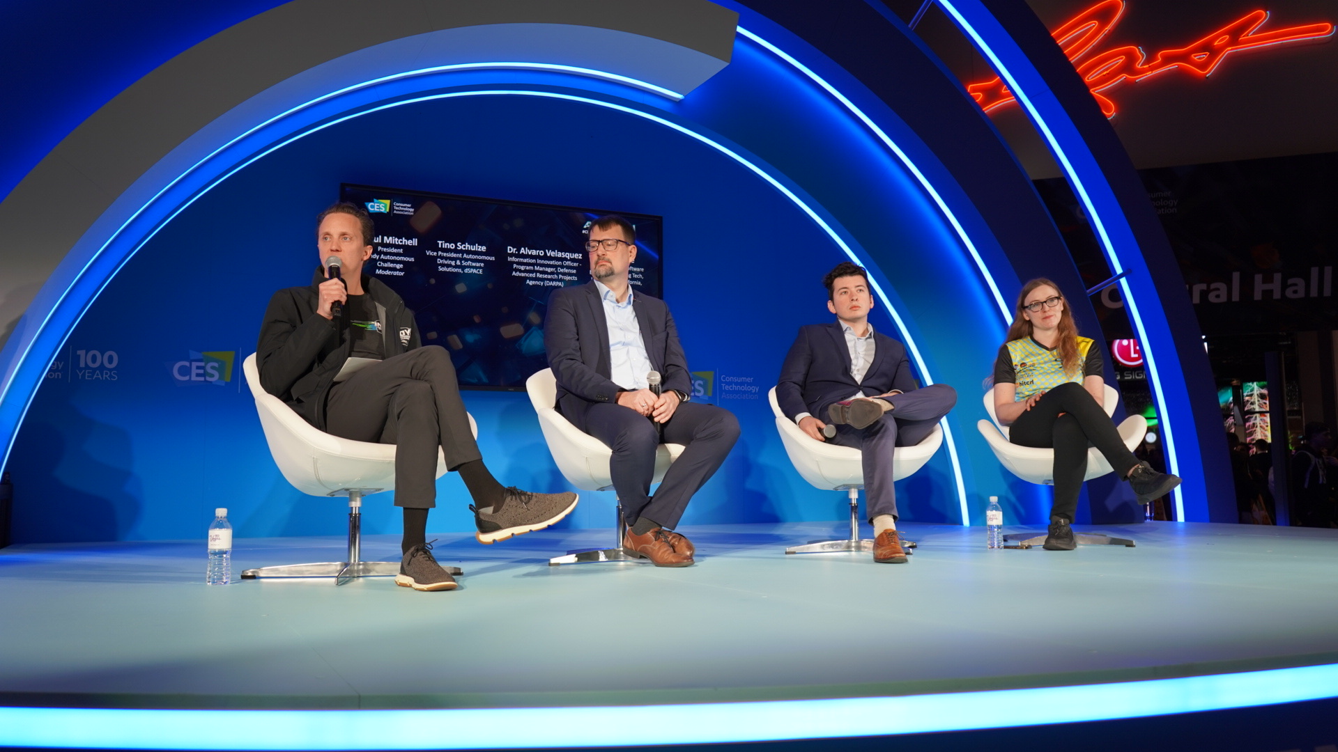 Four panelists seated in white swivel chairs on a blue-lit CES stage during a keynote discussion